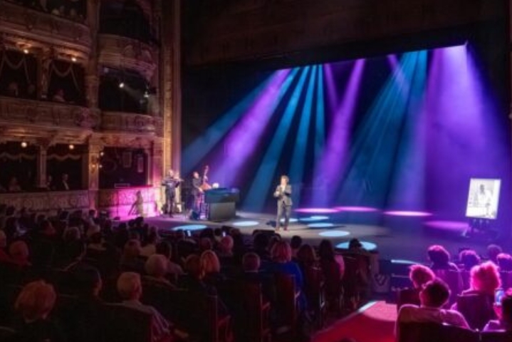Widownia siedzi w teatrze i ogląda wydarzenie na scenie. W centralnej części sceny stoi osoba trzymająca mikrofon. Po lewej stronie znajdują się muzycy z instrumentami. Nad sceną świecą fioletowe i niebieskie reflektory. Po prawej stronie stoi podświetlony portret w ramie. Po lewej stronie kadru widoczne są balkony teatru z publicznością. Na grafice nie ma widocznych napisów.
