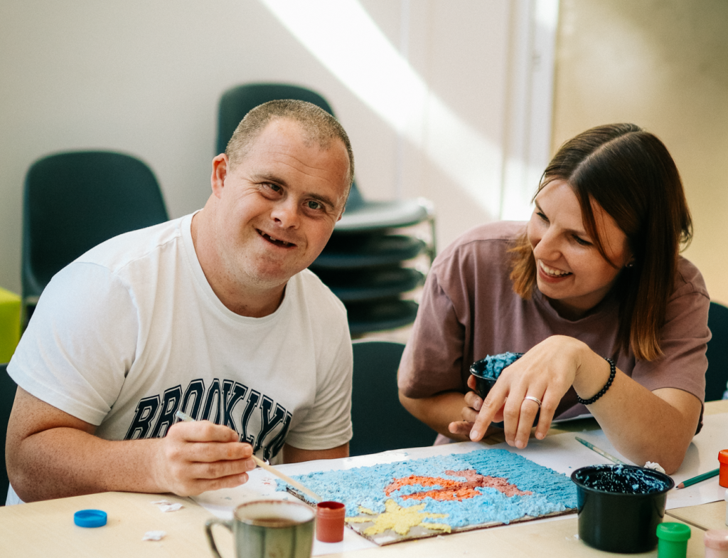 Dwie osoby siedzą przy stole z materiałami plastycznymi. Osoba po lewej ma krótkie włosy i jasną koszulkę z widocznym napisem Brooklyn. Osoba po prawej ma długie włosy i trzyma w dłoni narzędzie do tworzenia mozaiki. Na blacie leżą kolorowe elementy układanki.