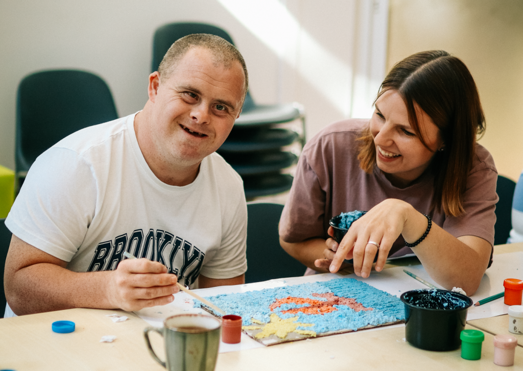 Dwie osoby siedzą przy stole w jasnym pomieszczeniu. Mężczyzna siedzi po lewej stronie. Ma krótkie włosy, patrzy w stronę obiektywu i trzyma pędzel. Nosi biały T-shirt z czarnym napisem BROOKLYN. Kobieta siedzi po prawej stronie. Ma średniej długości włosy i trzyma pojemnik z ciemną farbą. Oboje pracują nad kolorową pracą plastyczną ułożoną na stole. Przed nimi stoją otwarte pojemniki z farbami.