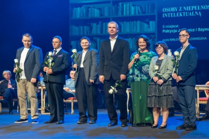 Na scenie stoi siedem osób ustawionych w rzędzie. Każda trzyma białą różę. W środku stoi wysoki mężczyzna w ciemnym garniturze. Obok niego stoi kobieta z ciemnymi włosami w zielonej bluzce i zielonej spódnicy. Pozostałe osoby mają garnitury lub eleganckie stroje. W tle siedzą uczestnicy wydarzenia. Na ekranie po prawej stronie widoczne są napisy: osoby z niepełnosprawnością intelektualną, II miejsce: Patrycja Jarychowicz i Szymon oraz czyta Małgorzata Zawadzka. Część tekstu z nazwiskami jest poza kadrem i pozostaje nieczytelna.