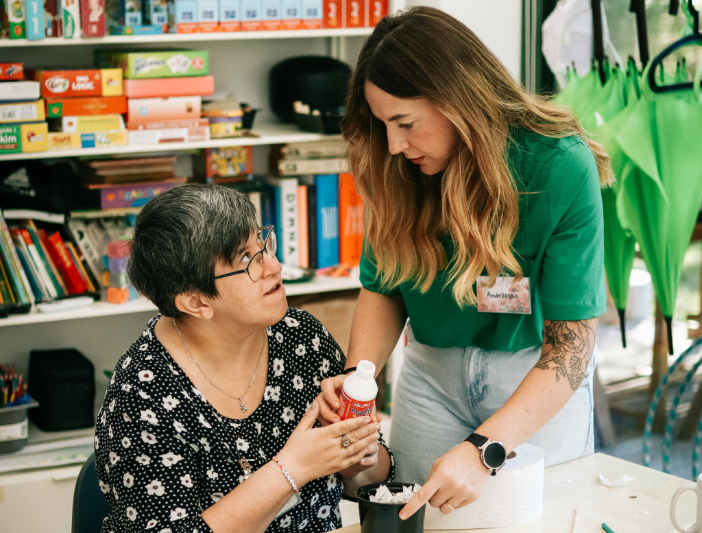 Dwie osoby siedzą przy stole w pomieszczeniu z półkami pełnymi książek i materiałów. Osoba po lewej ma krótkie, ciemne włosy, okulary i patrzy na drugą osobę. Osoba po prawej ma długie, jasne włosy i pochyla się nad stołem, trzymając dłoń drugiej osoby.