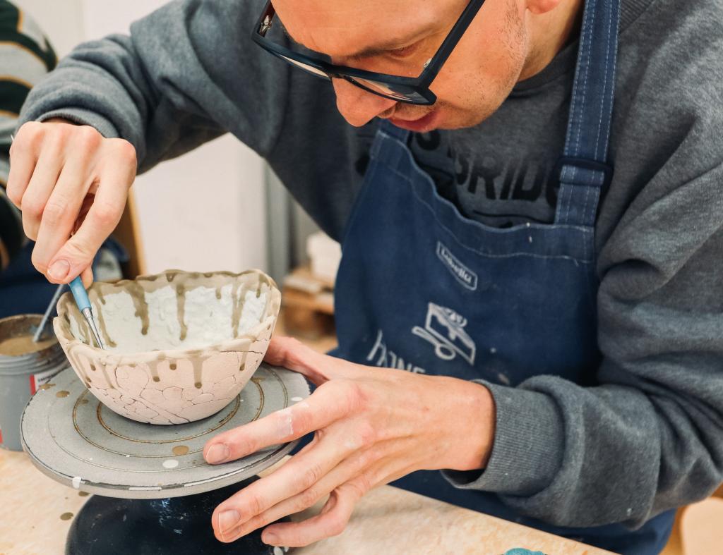 Mężczyzna w okularach i granatowym fartuchu siedzi przy stole. Jego dłonie trzymają jasną ceramiczną miseczkę z brązowymi zaciekami, która stoi na toczku. Prawą ręką nanosi brązowe szkliwo pędzelkiem na miseczkę. Na fartuchu widnieje napis: LUBELLA. Obok toczka stoi słoiczek ze szkliwem i pędzlem. W tle widać inną osobę, fragment blatu i ścianę.