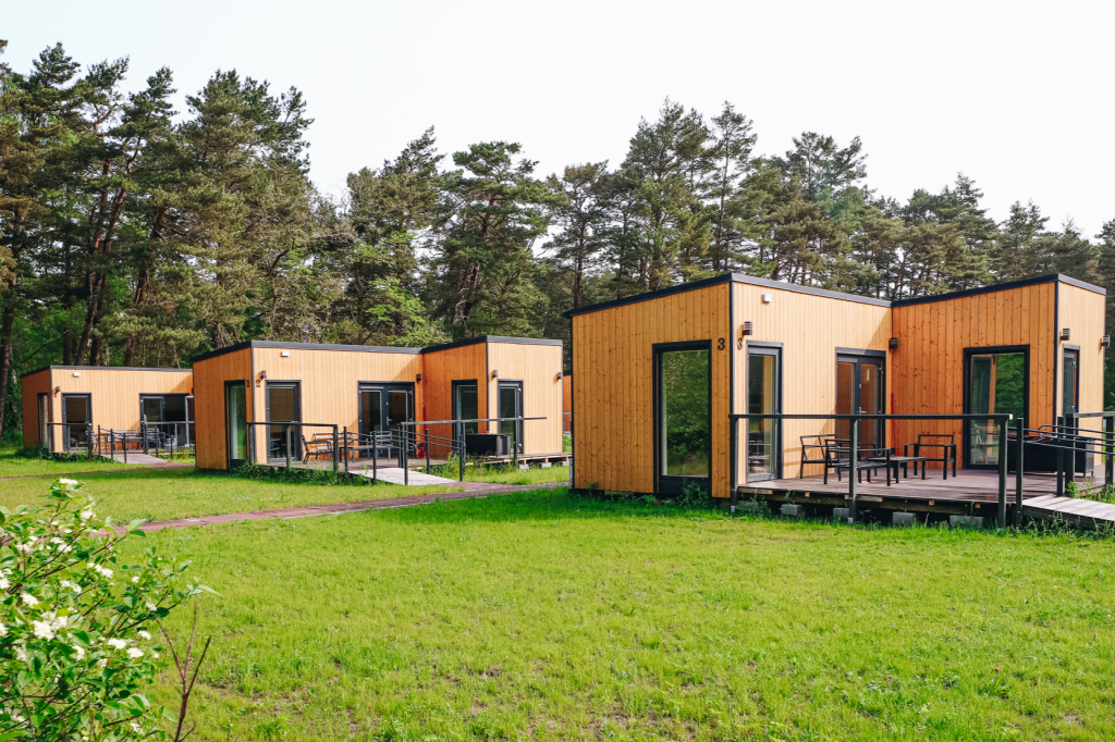 W pierwszym planie znajduje się trawnik. W dalszym planie widać parterowe budynki w jasnych i ciemnych kolorach, otoczone drzewami.